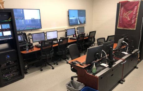 The SmartField Control Room used by Boston College Football is in a beige collored room. Computer monitors and joystick camera controllers and switchers sit atop desks with monitors. Two monitors are also mounted on the wall. The room is used to control all cameras used at the BC football practice facility. The footage will be used by BC coaches to work with athletes and assess player development.