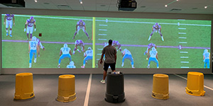 A player stands at a 9' tall wall, matching up with players from the opposing team projected lifesize onto the wall.