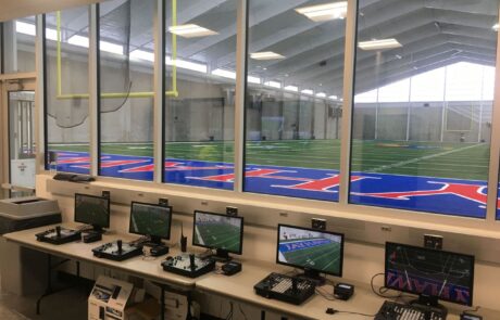 Photo of the Kansas University football video command and control center with 5 displays and joystick controllers for remote-control cameras in their indoor practice facility, which is visible through the wall of windows above the desk of video displays and controllers.
