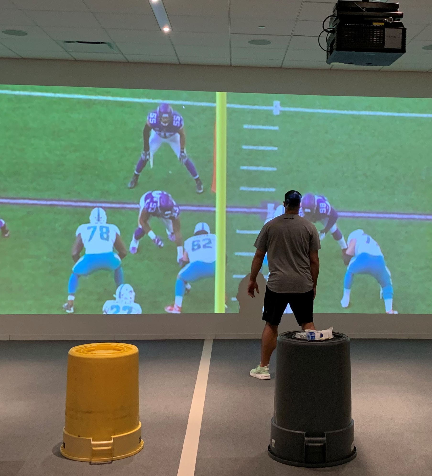 A NY Jets player stands in front of a full-wall video screen with an end zone view of an American football play, showing that the size of the players on the screen matches the size of the player.