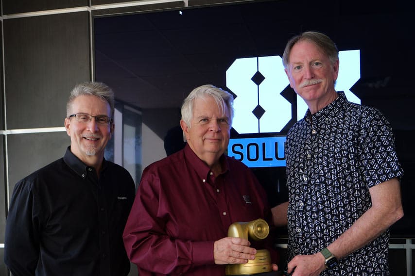 Wes Carr of Panasonic, Dan Aton of 8K Solutions, and Pat Lenihan of Panasolic stand in front of a digital screen with the 8K Solutions logo. Dan is holding the Golden PTZ Award presented to 8K Solutions for innovation using Panasonic pan-tilt-zoom cameras.