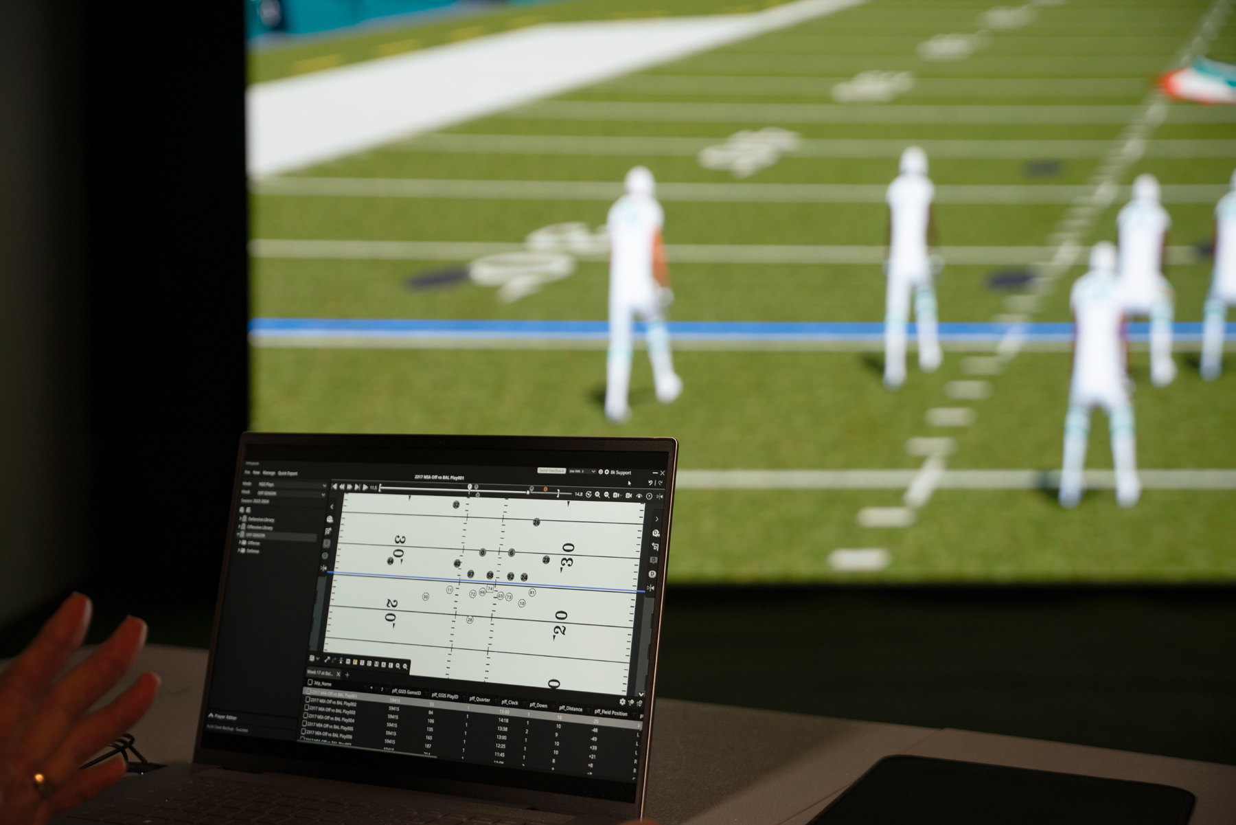 Looking over the shoulder of a coach customizing formations using 3D Playbook on a laptop to see the animated formation on a lifesize Walk Through Wall. The formation appears as Xs, Os, lines, and arrows on the computer game, but a motion animation on the lifesize screen