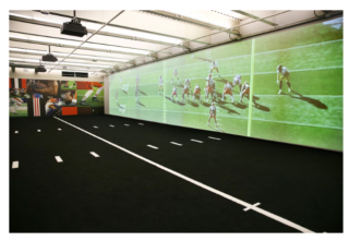 A 25-foot wide 8K Solutions Walk Through Wall with a game replay on the screen is shown on the third floor of the Browns' new weight room. The floor is covered in brown astroturf.