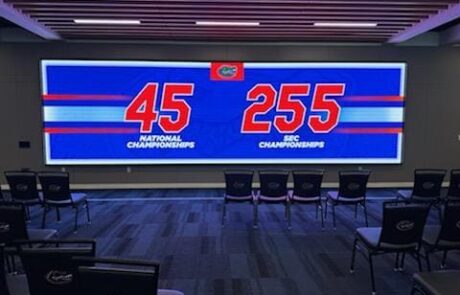 Image of the LED Walk Through Wall at the University of Florida Gator Football Complex. In the darkened room, the 30' wide by 10' high wall displays records earned by the team. A few croups of chairs face the wall.